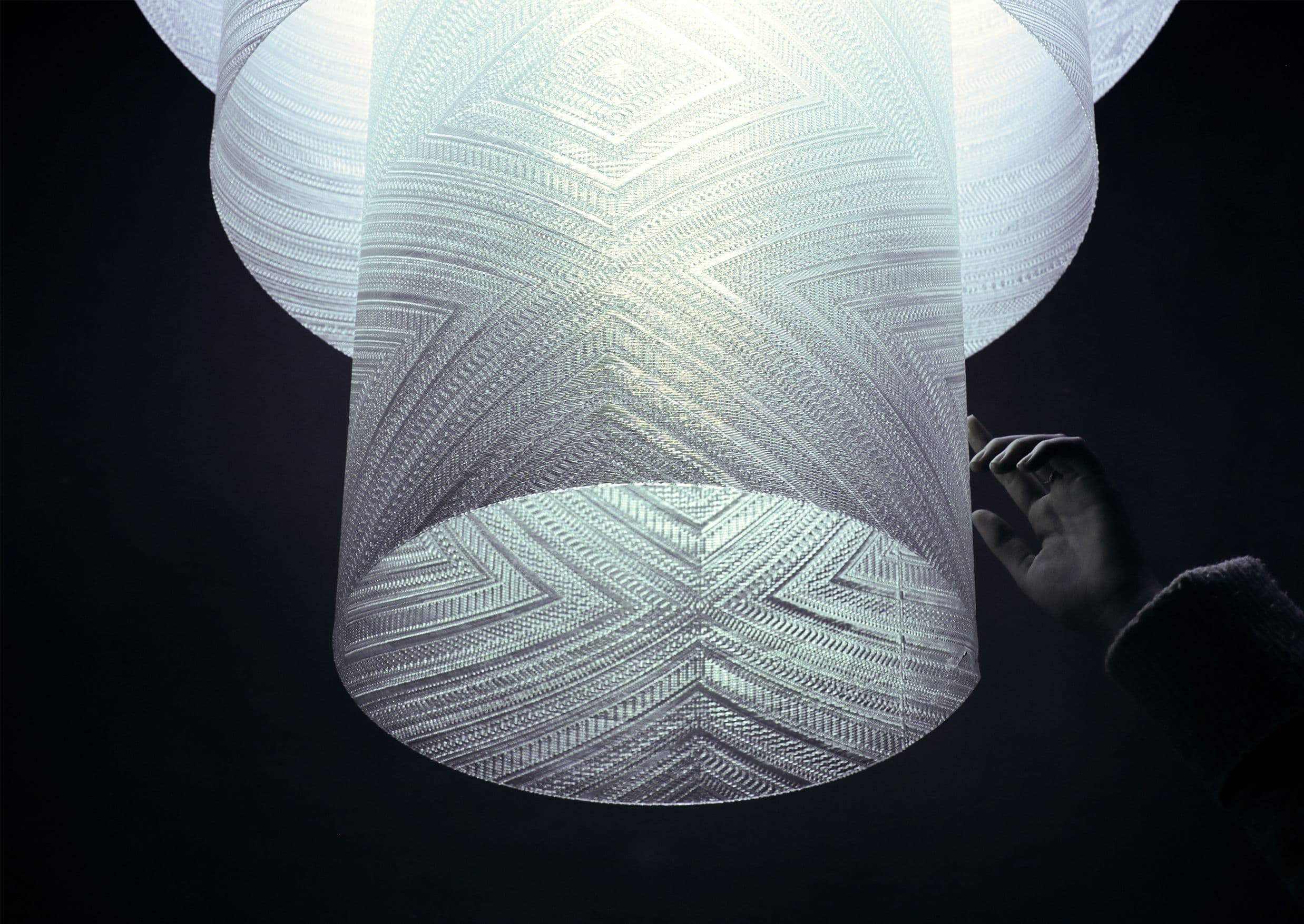 image of a textured light shade with a black background, a hand is reaching up to touch the light shade