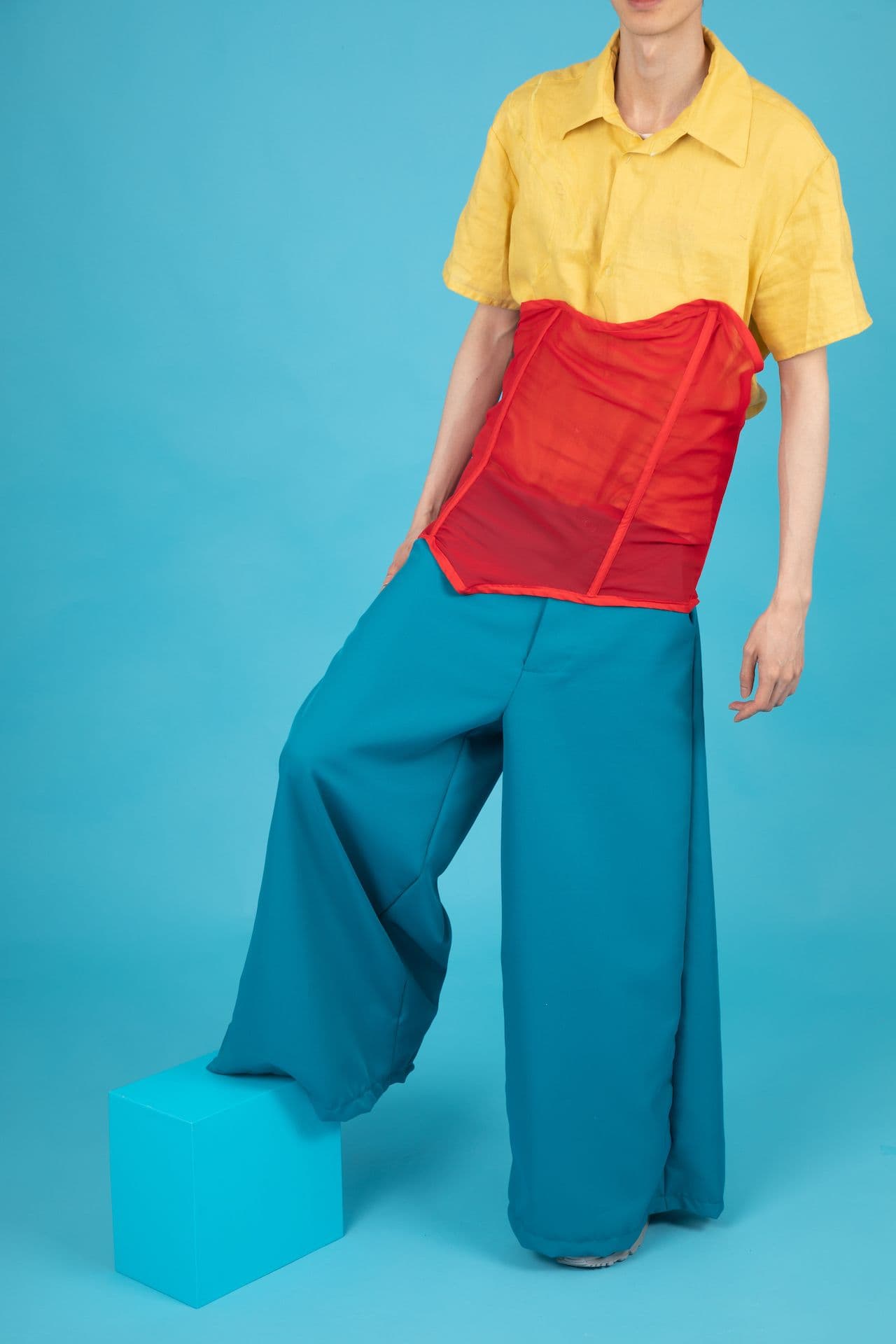 Person standing in a blue room wearing blue pants, red corset and yellow shirt.